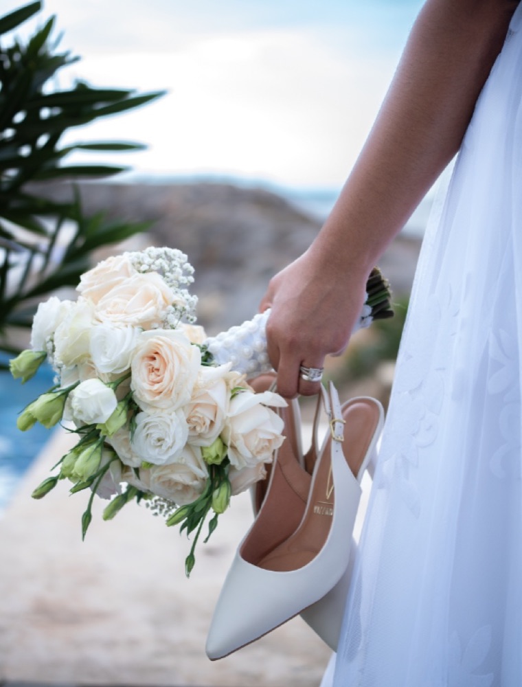 Novia con ramo de rosas blancas y zapatos en la mano frente al mar